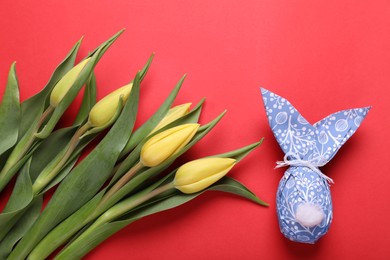 Photo of Easter bunny made of wrapping paper and egg near beautiful tulips on red background, flat lay