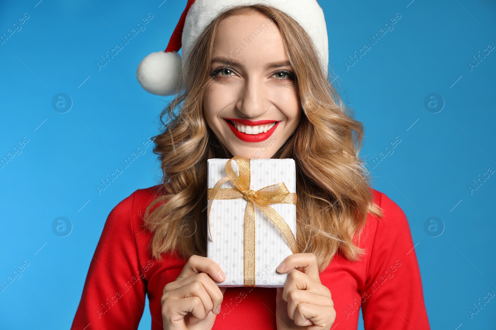 Photo of Happy young woman wearing Santa hat with Christmas gift on light blue background, closeup