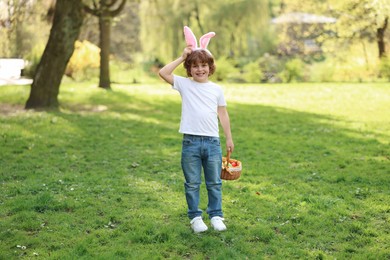 Photo of Easter celebration. Cute little boy in bunny ears holding wicker basket with painted eggs outdoors