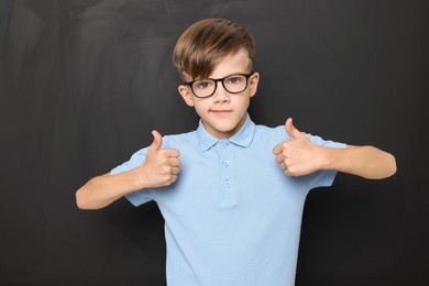 Photo of Cute schoolboy in glasses showing thumbs up near chalkboard