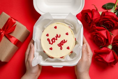 Photo of Woman holding takeaway box with bento cake at red table, top view. St. Valentine's day surprise