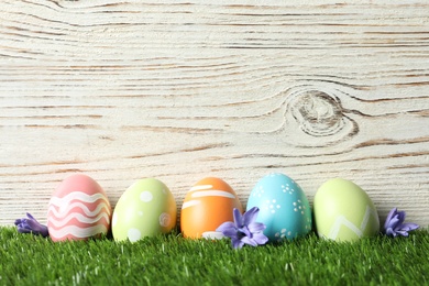 Colorful painted Easter eggs with flowers on green grass against wooden background, space for text