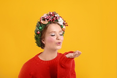 Beautiful young woman wearing Christmas wreath on yellow background