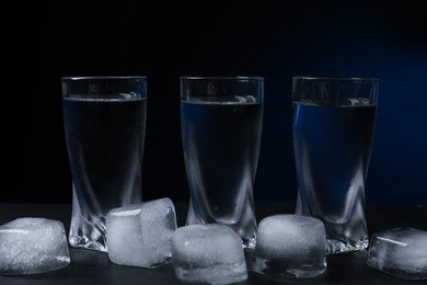 Photo of Shot glasses of vodka with ice cubes on black table against dark background