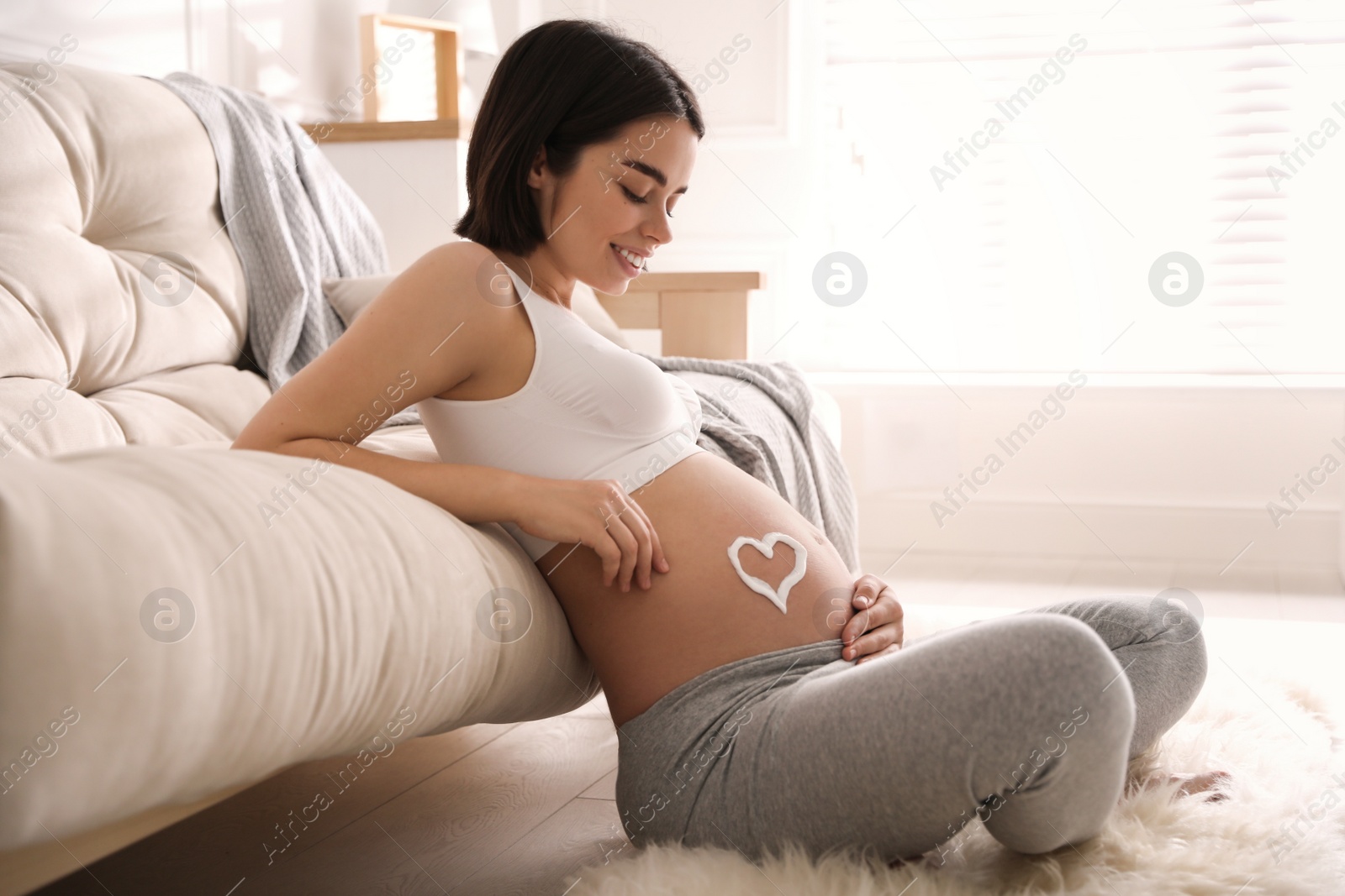 Photo of Young pregnant woman with cosmetic product on belly at home