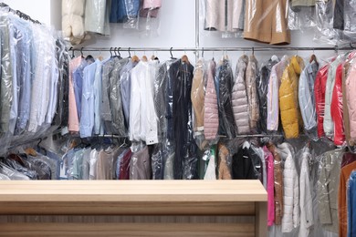 Photo of Dry-cleaning service. Hangers with different clothes in plastic bags on racks and wooden counter indoors