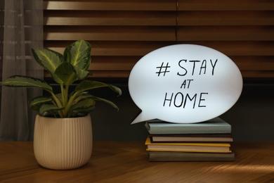 Photo of Houseplant, books and speech bubble with hashtag STAY AT HOME on wooden table. Message to promote self-isolation during COVID‑19