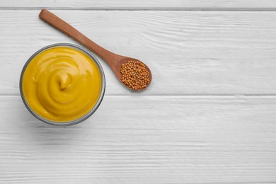 Photo of Bowl of delicious mustard and spoon with seeds on white wooden table, flat lay. Space for text