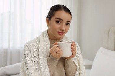 Photo of Beautiful young woman with cup of drink wrapped in knitted plaid at home