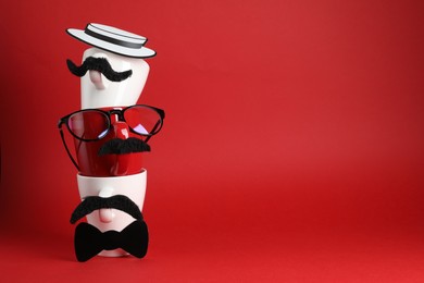 Photo of Men's faces made of cups, fake mustaches, glasses, hat and bow tie on red background. Space for text