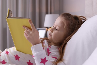 Photo of Cute little girl reading book on bed at home