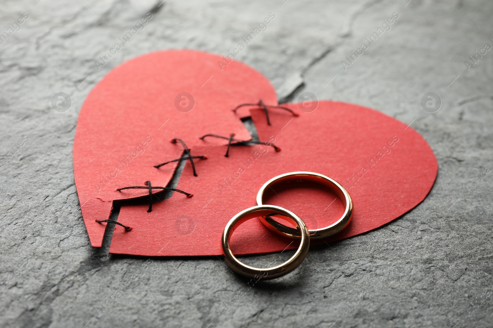 Photo of Broken heart. Torn red paper heart sewed with thread and wedding rings on grey table, closeup