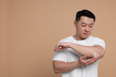 Handsome man applying body cream onto his elbow on light brown background. Space for text