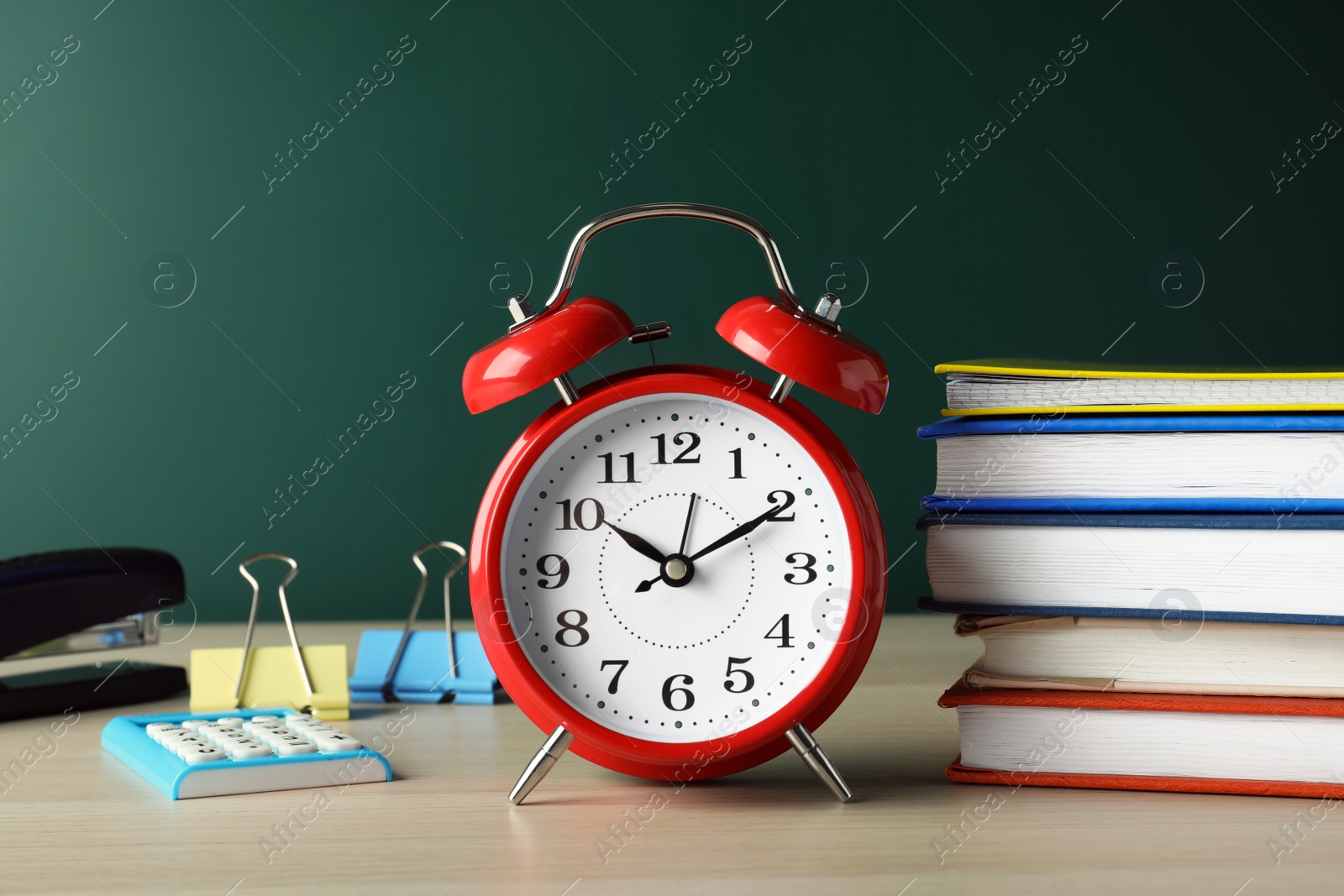 Photo of Alarm clock and different stationery on wooden table near green chalkboard. School time