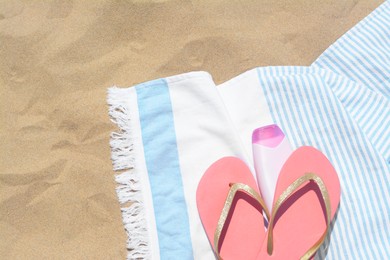 Striped towel with bottle of sunblock and flip flops on sandy beach, flat lay. Space for text