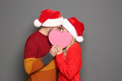 Young couple in Santa hats hiding behind heart on grey background. Christmas celebration
