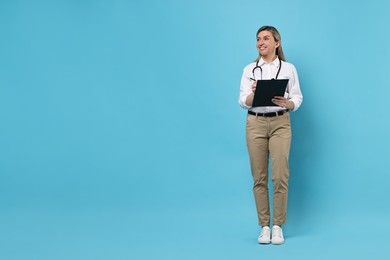 Portrait of happy doctor with stethoscope and clipboard on light blue background, space for text