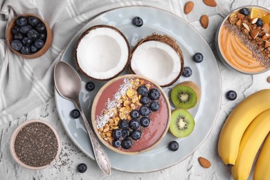 Photo of Bowl of delicious fruit smoothie with fresh blueberries, granola and different ingredients on white textured table, flat lay