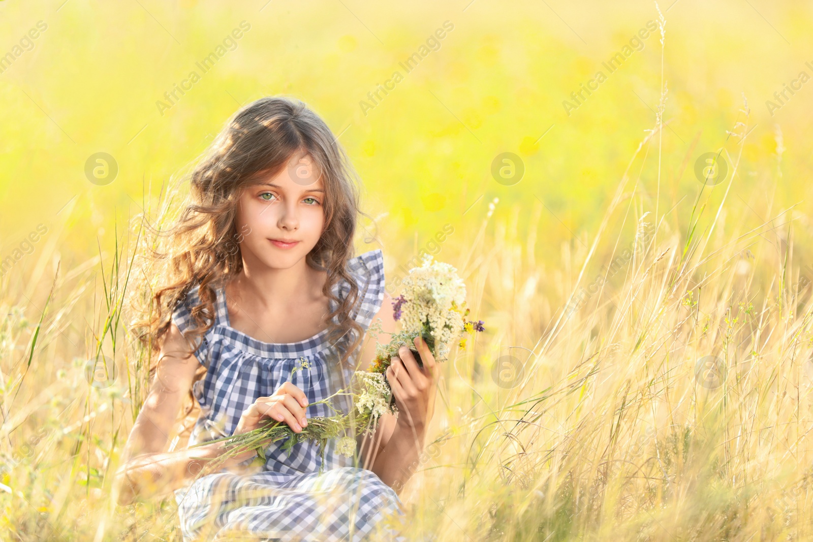 Photo of Cute little girl making wreath of beautiful flowers in field on sunny day