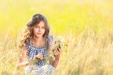 Photo of Cute little girl making wreath of beautiful flowers in field on sunny day