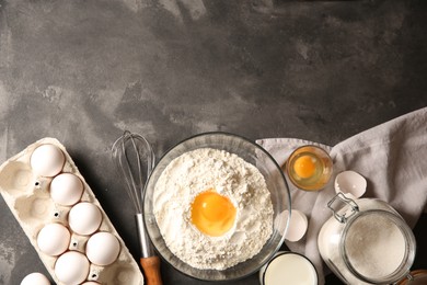 Photo of Making dough. Flour with egg yolk in bowl and other products on grey textured table, flat lay. Space for text