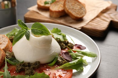 Photo of Delicious burrata salad with tomatoes, arugula and pesto sauce on dark grey table, closeup