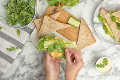 Photo of Woman adding arugula to tasty sandwich at white marble table, top view