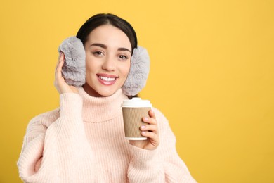 Photo of Beautiful young woman in earmuffs with cup of drink on yellow background. Space for text