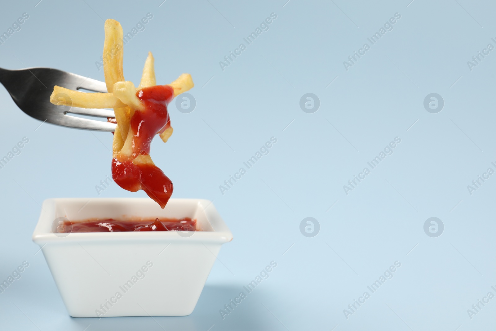 Photo of Dipping delicious french fries into ketchup on light blue background, closeup. Space for text