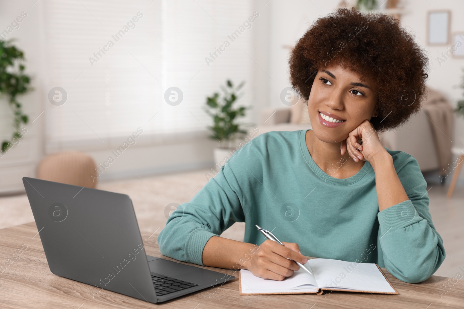 Photo of Young woman using laptop and writing in notebook at wooden desk in room