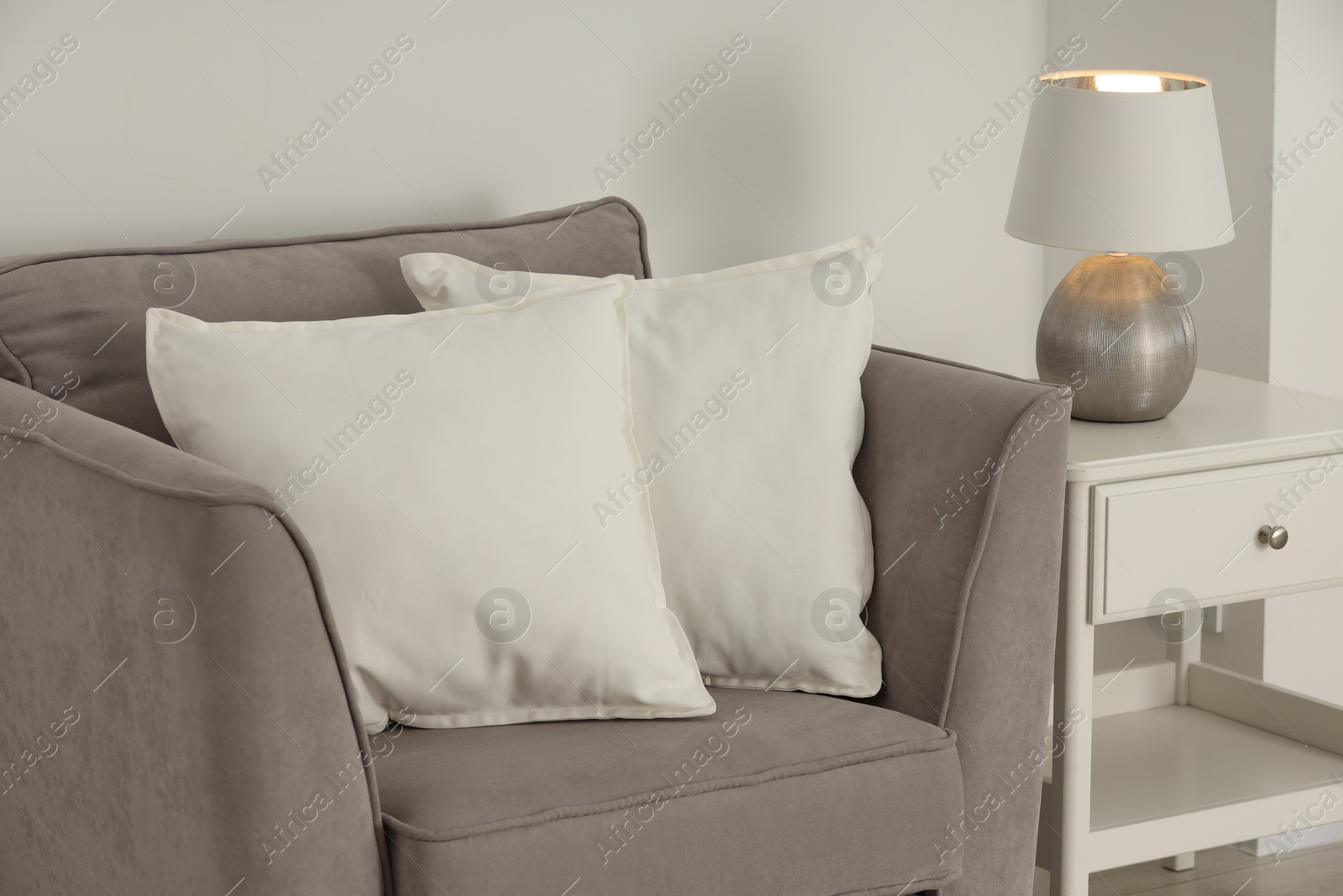 Photo of Soft white pillows on armchair, bedside table and lamp near light wall indoors