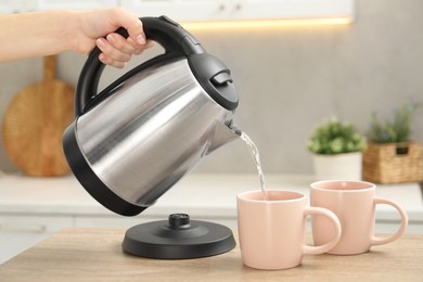 Photo of Woman pouring hot water from electric kettle into cup in kitchen, closeup