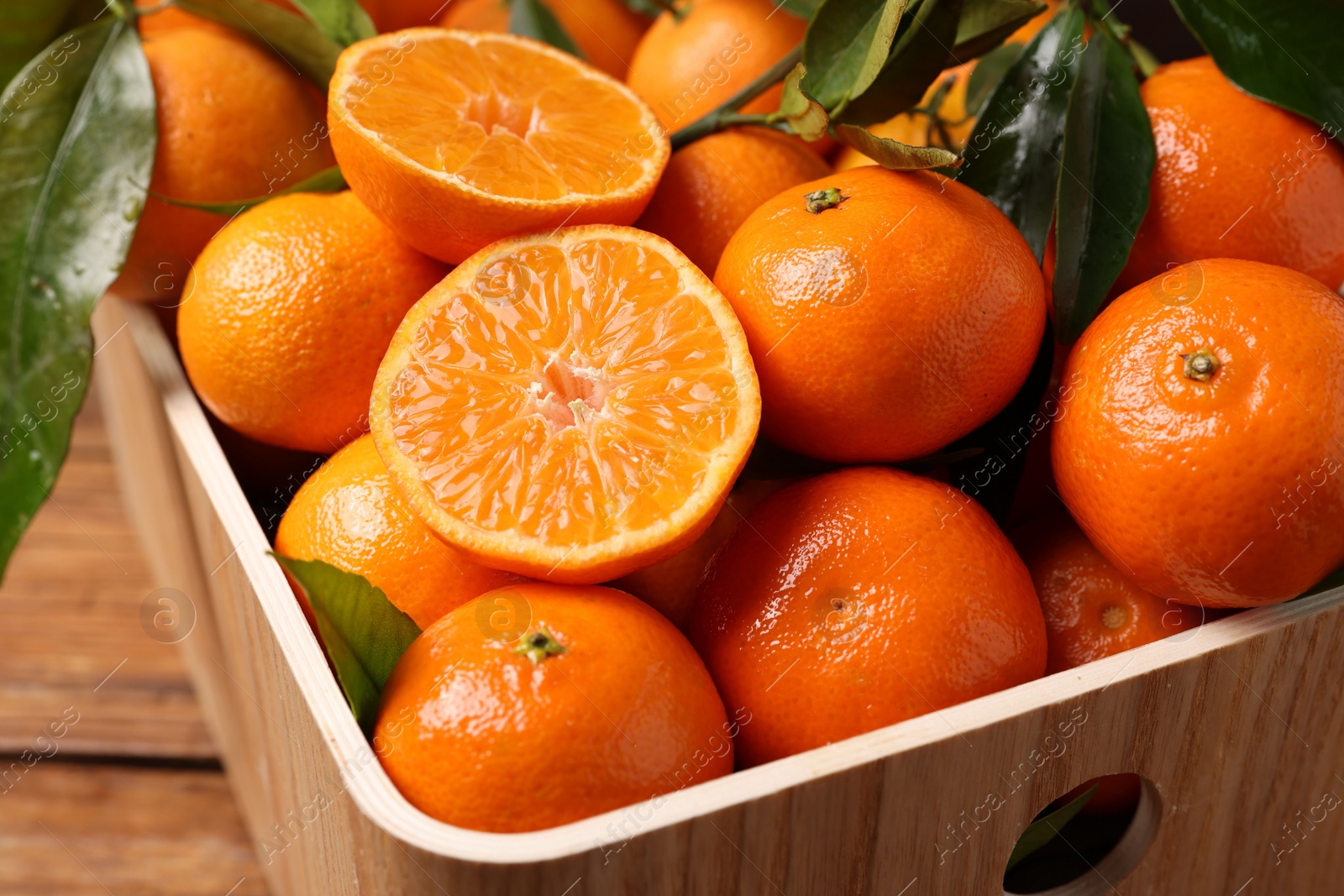 Photo of Fresh tangerines with green leaves in crate on wooden table, closeup