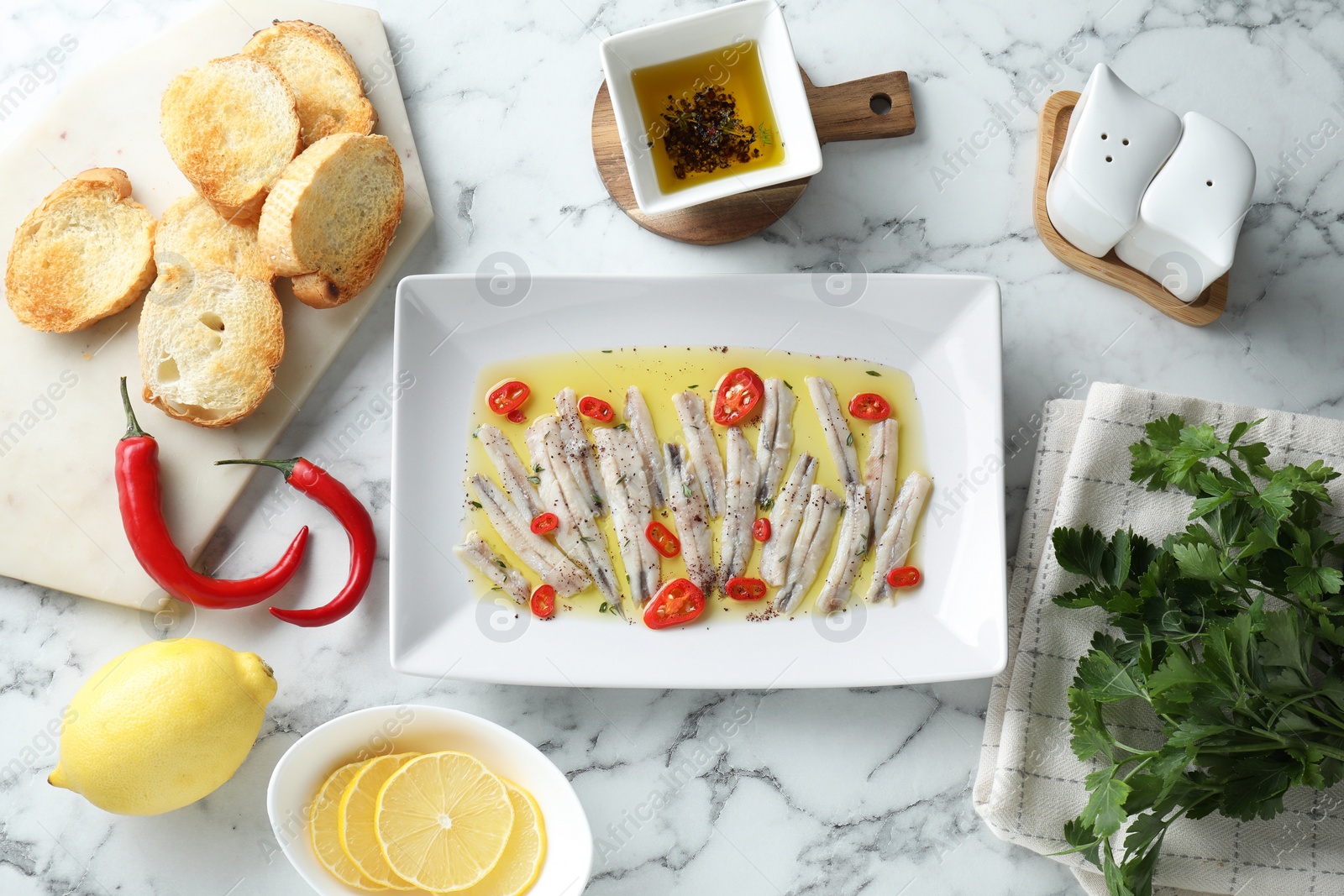 Photo of Tasty pickled anchovies with spices and products on white marble table, flat lay