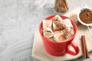 Photo of Cup of aromatic hot chocolate with marshmallows, cocoa powder and cinnamon sticks on gray table, closeup. Space for text