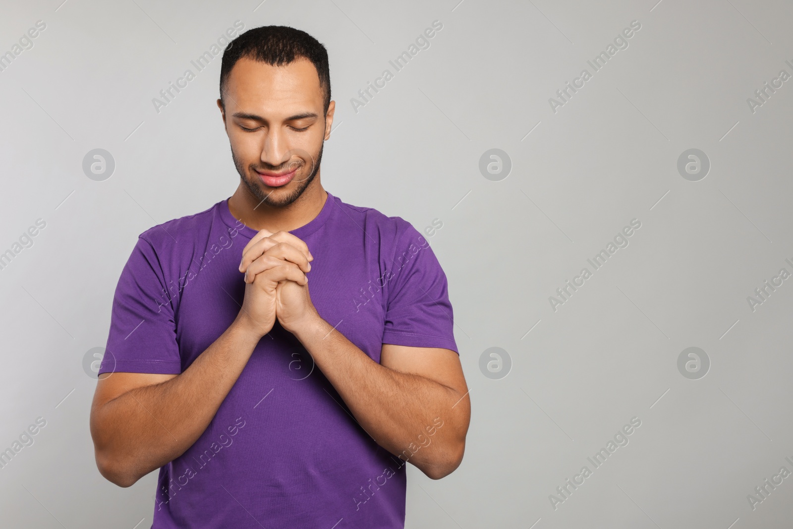 Photo of African American man with clasped hands praying to God on light grey background. Space for text