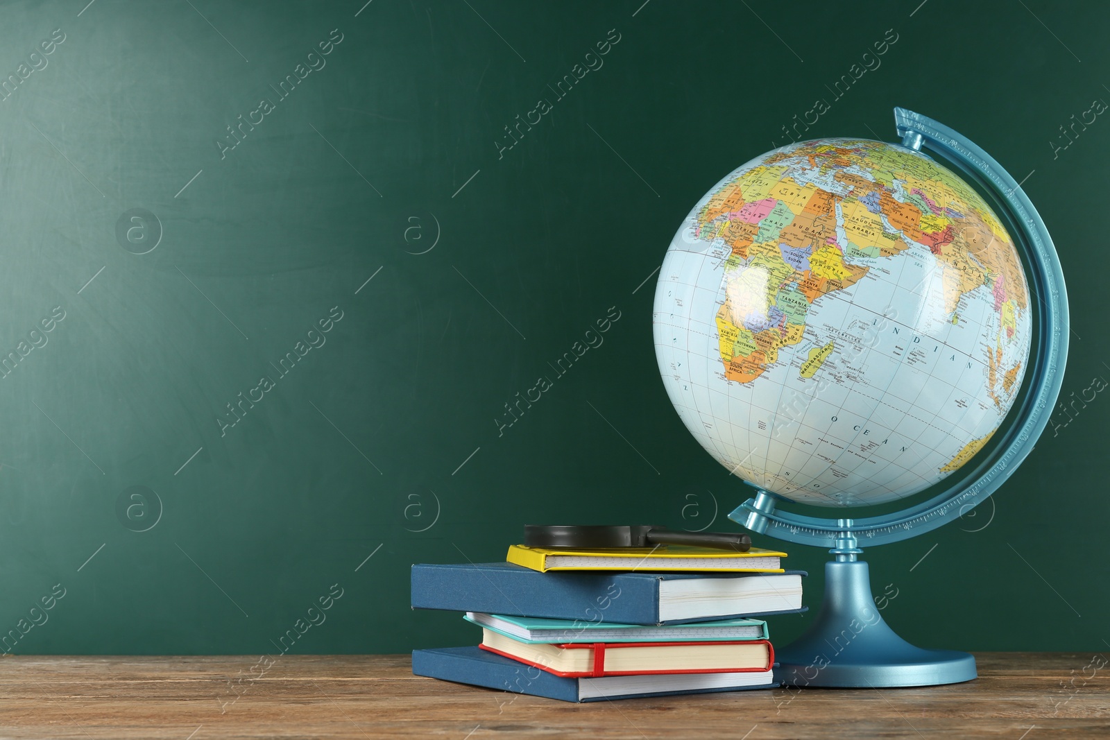 Photo of Globe, books and magnifying glass on wooden table near green chalkboard, space for text. Geography lesson