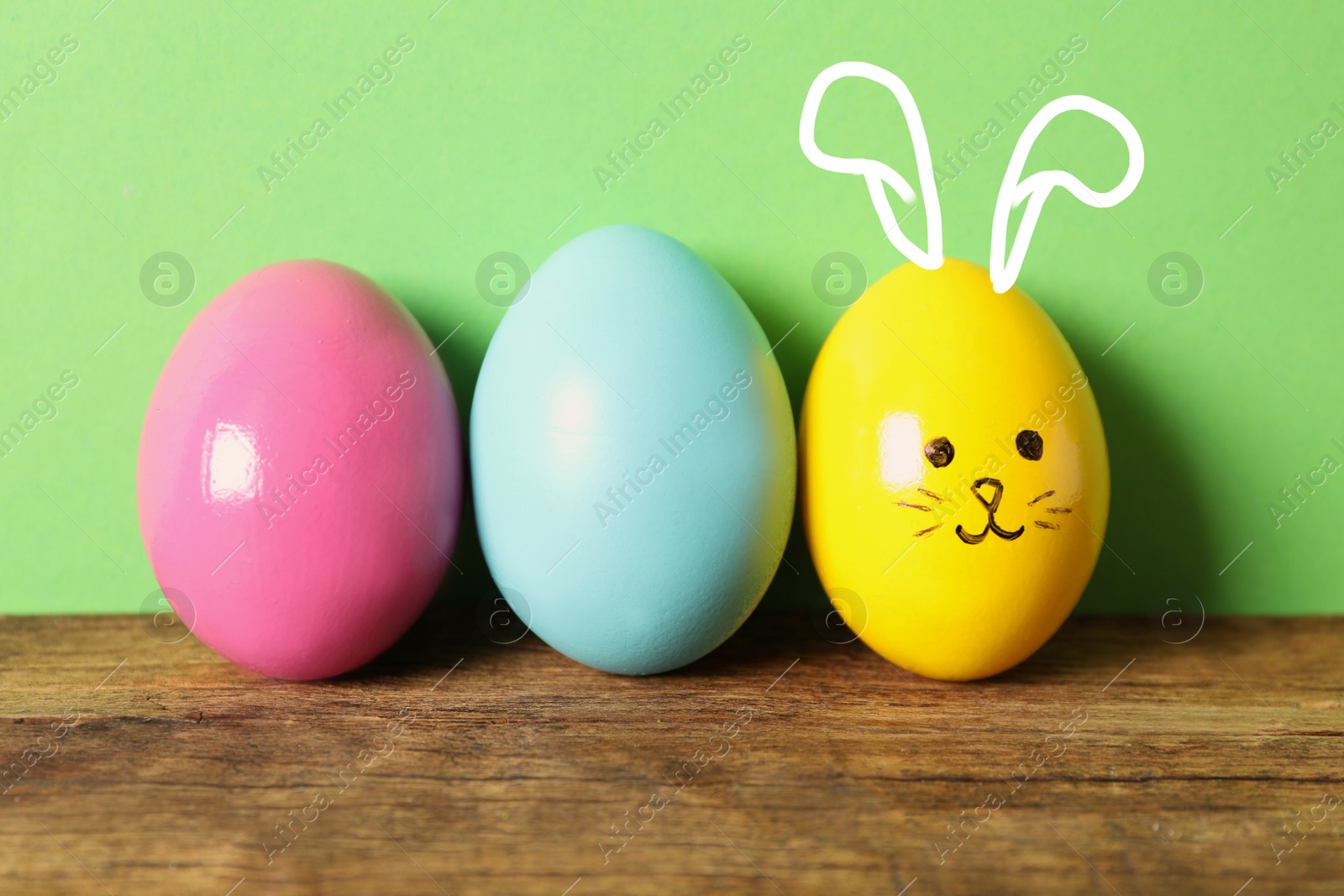 Image of Yellow egg with drawn face and ears as Easter bunny among others on wooden table against green background