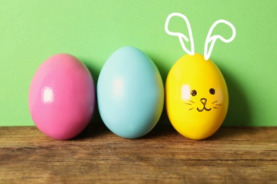 Yellow egg with drawn face and ears as Easter bunny among others on wooden table against green background
