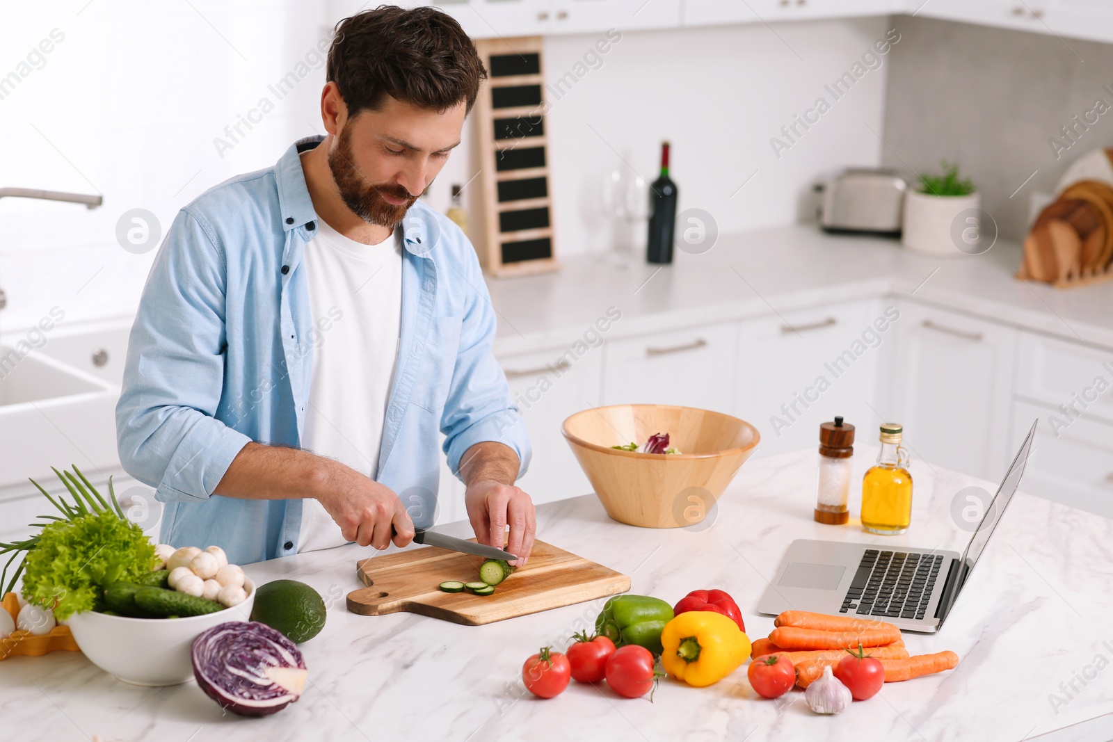 Photo of Man making dinner while watching online cooking course via laptop in kitchen