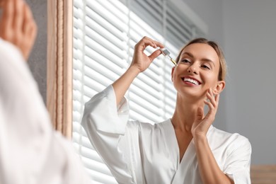 Beautiful woman applying cosmetic serum onto her face near mirror in bathroom