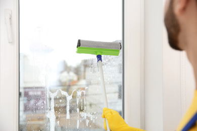 Photo of Professional young janitor in uniform cleaning window indoors, closeup
