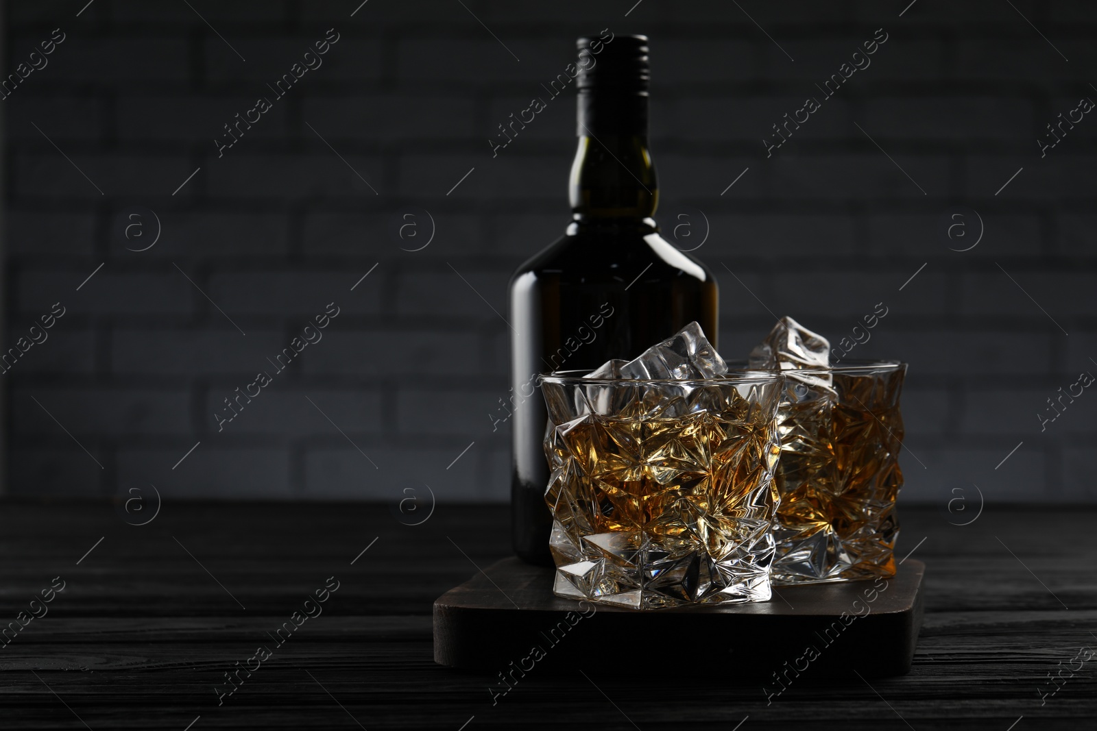 Photo of Whiskey with ice cubes in glasses and bottle on black wooden table, space for text