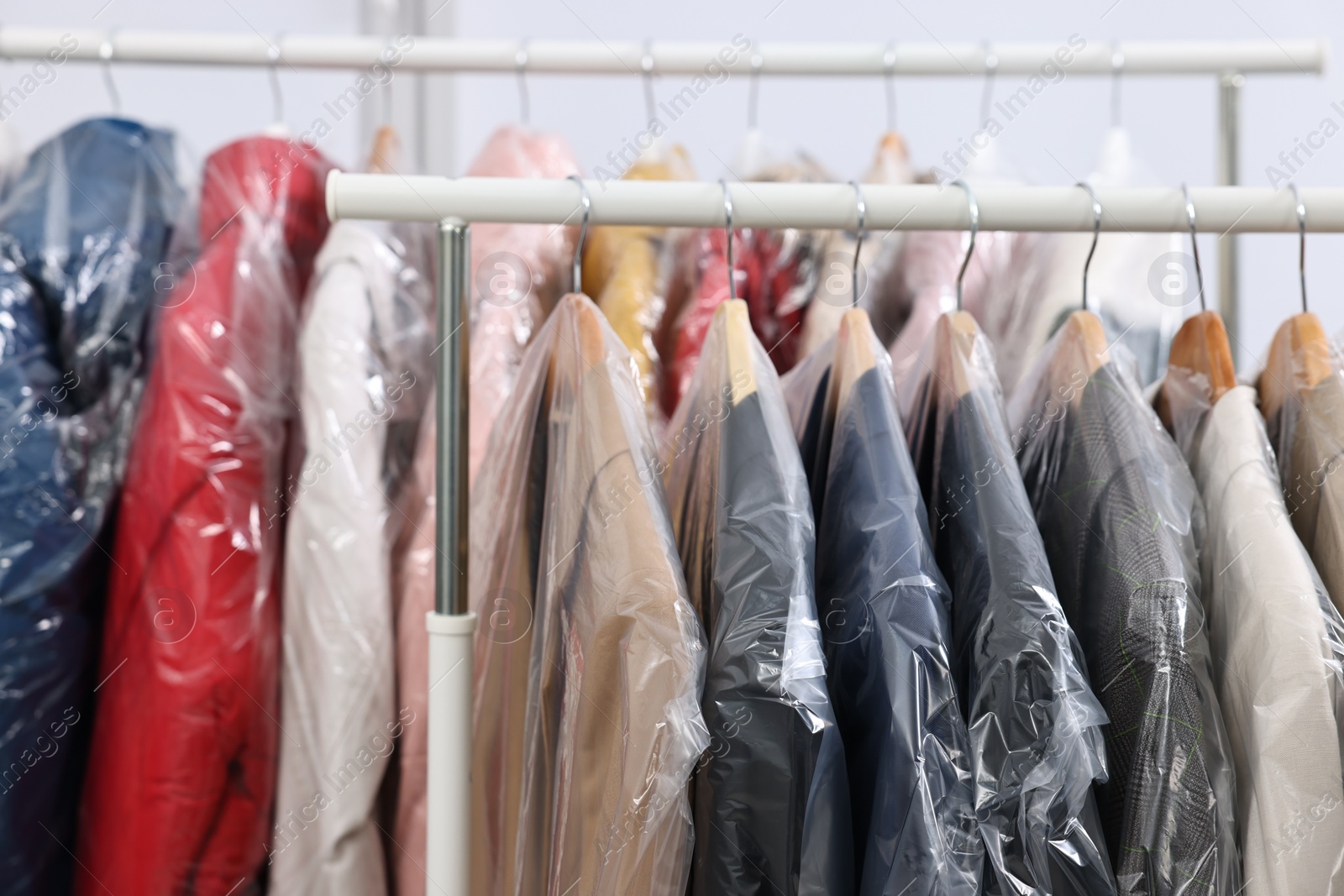 Photo of Dry-cleaning service. Many different clothes in plastic bags hanging on rack indoors, closeup