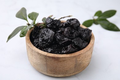 Sweet dried prunes in bowl and green leaves on white table, closeup
