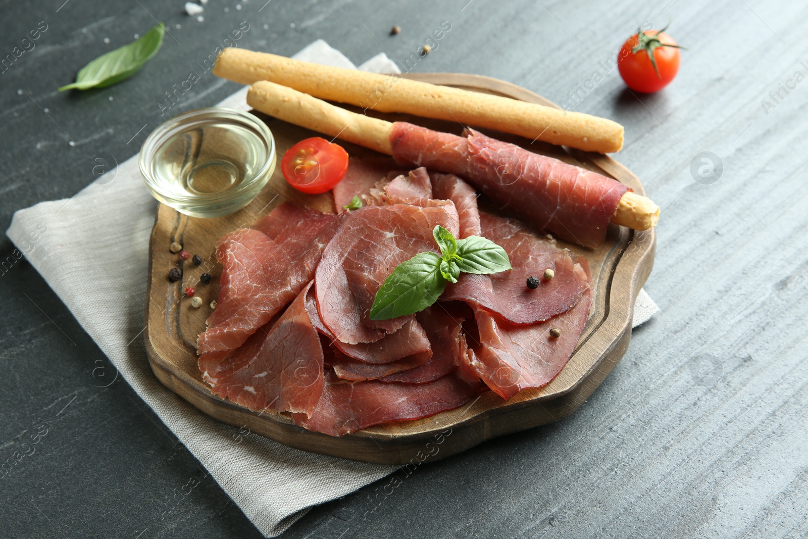 Photo of Delicious bresaola, tomato, grissini sticks and basil leaves on grey textured table