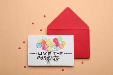 Photo of Card with phrase Live The Moments and red envelope on beige background, flat lay