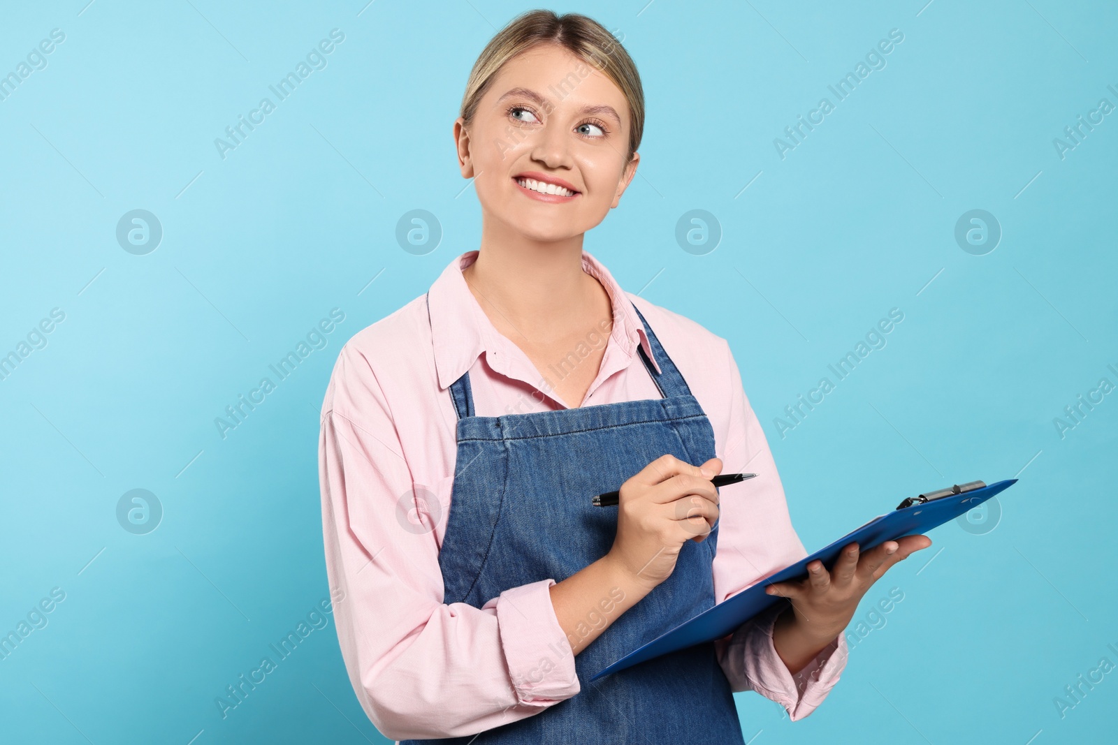 Photo of Beautiful young woman in denim apron with clipboard on light blue background. Space for text