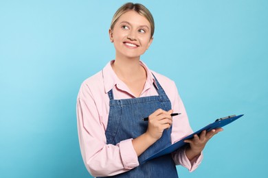 Photo of Beautiful young woman in denim apron with clipboard on light blue background. Space for text