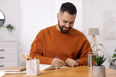 Young man writing in notebook at wooden table indoors